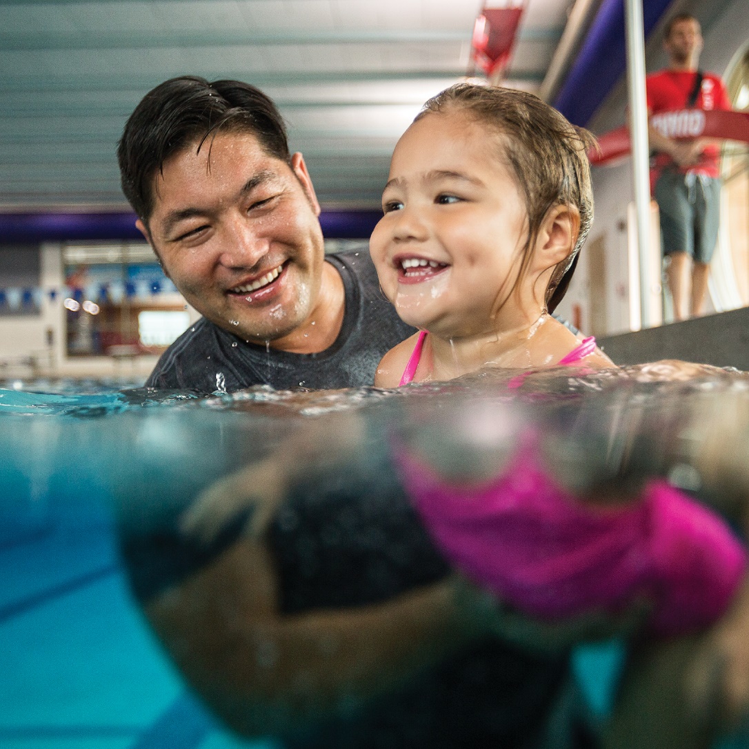 A joyful child in a pink swimsuit learns to swim with the guidance of a smiling adult in an indoor pool, with a lifeguard overseeing in the background.