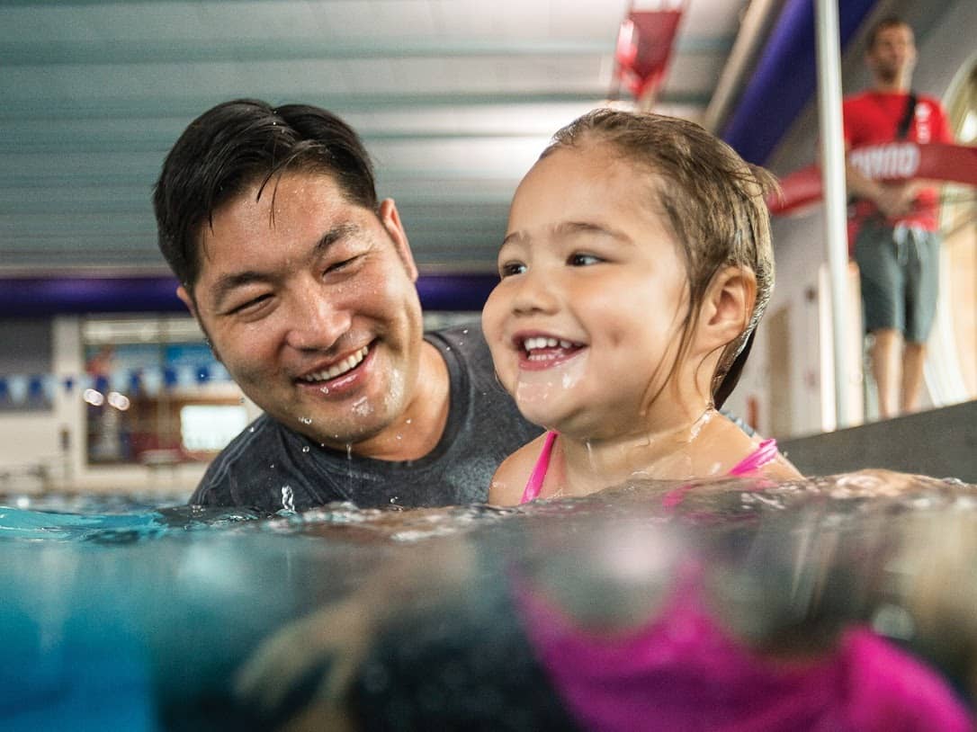 A smiling man and young girl enjoying a swimming lesson at the Owen County YMCA pool. The girl is wearing a pink swimsuit, and the man is partially submerged, engaging with her as they share a joyful moment in the water.